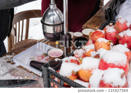 Fresh pomegranates covered with snow sit beside manual juice press, paper cups at outdoor stand, vivid red fruit contrasts with metal tools 135927369