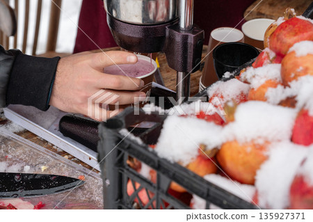 Fresh pomegranate juice is poured into a paper cup at street stand, bright red fruit covered with snow contrasts with metal press and winter textures 135927371