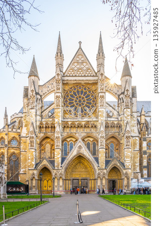 People gather near Westminster Abbey in London during the early morning. The north front shows detailed architecture and the large rose window. Trees frame the scene in a clear sky. People gather near Westminster Abbey in London during the early morning. The north front shows detailed architecture and the large rose window. Trees frame the scene in a clear sky. 135927485