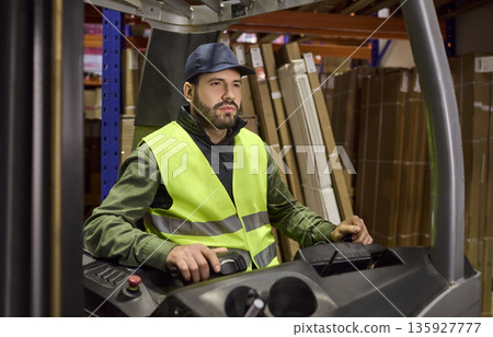 Warehouse forklift operator in safety vest driving loader among shelving Warehouse forklift operator in safety vest driving loader among shelving 135927777