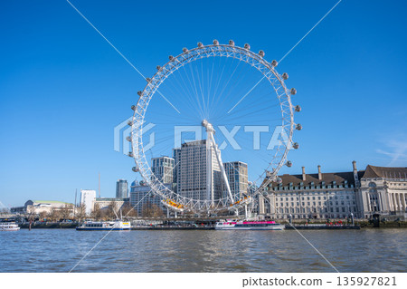 The London Eye towers above the Thames River. People stroll along the riverbank as boats travel. Buildings surround the area under clear blue skies. The London Eye towers above the Thames River. People stroll along the riverbank as boats travel. Buildings surround the area under clear blue skies. 135927821
