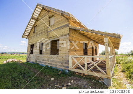 Wide angle view of unfinished wooden ecological traditional cottage of natural lumber materials with steep roof and porch under construction in green neighborhood on blue sky copy space background. 135928268