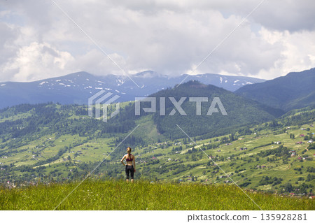 Back view of slim young woman standing on grassy valley on background of green mountains on sunny summer day. 135928281