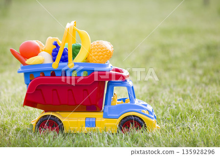 Bright plastic colorful toy car truck carrying basket with toy fruits and vegetables outdoors on sunny summer day. Bright plastic colorful toy car truck carrying basket with toy fruits and vegetables outdoors on sunny summer day. 135928296