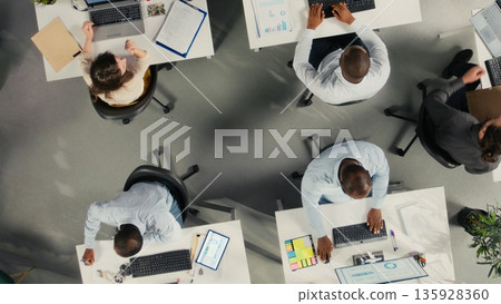 Top down view of employees consulting on new corporate project with charts in workspace, staff aligning targets with performance metrics for resource allocation and reports. 135928360