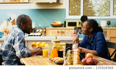 African american couple serving coffee in mugs and enjoying weekend morning together, bonding and relaxing at the breakfast table. Young happy people sharing a positive conversation. African american couple serving coffee in mugs and enjoying weekend morning together, bonding and relaxing at the breakfast table. Young happy people sharing a positive conversation. 135928369