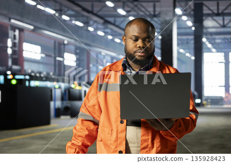 Black industrial personnel overseeing workshop fabrication equipment on laptop, emphasizing heavy industry safety and advanced development with automation in the manufacturing process. Black industrial personnel overseeing workshop fabrication equipment on laptop, emphasizing heavy industry safety and advanced development with automation in the manufacturing process. 135928423