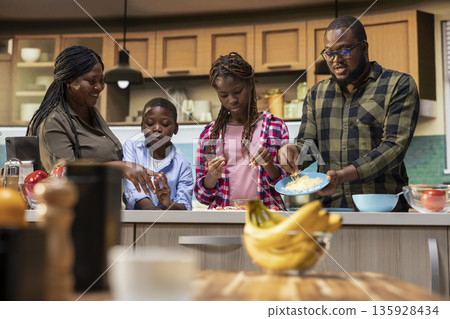 Black young kids and their parents adding grated mozzarella and cheese on dough, sharing pizza day at home and learning to cook with their parents for bonding time. Togetherness and laughter. 135928434