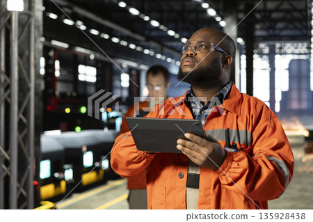 Black engineering supervisor in protective gear overseeing welding activity with tablet, reflecting heavy labor and the automation development of the industrial factory as sparks rise. Black engineering supervisor in protective gear overseeing welding activity with tablet, reflecting heavy labor and the automation development of the industrial factory as sparks rise. 135928438