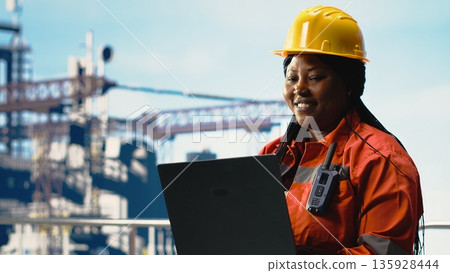 Portrait of cheerful offshore platform engineer managing supply operations with laptop. Happy oil rig technician records safety checks and inspection reports with notebook Portrait of cheerful offshore platform engineer managing supply operations with laptop. Happy oil rig technician records safety checks and inspection reports with notebook 135928444