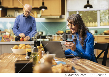 Elderly woman doing online shopping with a credit card in hand, reviewing list of groceries and daily essentials in e-commerce app cart on laptop. Female shopper checks credentials for order. 135928454