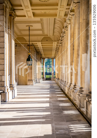 People walk through the corridor at Greenwich University in London during the day. Sunlight shines on the columns and floor, casting long shadows. The green landscape is seen outside. 135929006