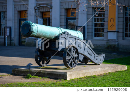 The historical cannon stands at the Old Royal Naval College in Greenwich, London. It is made of metal and sits on a stone base in front of a large building. 135929008