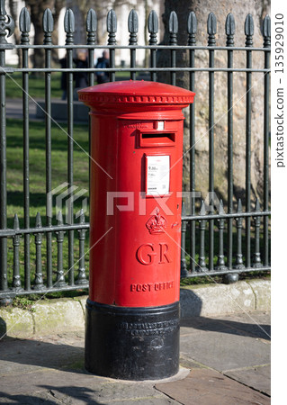 A traditional red post box is positioned on the street in London. It is surrounded by a metal fence and greenery. People can send letters and parcels from this post box. A traditional red post box is positioned on the street in London. It is surrounded by a metal fence and greenery. People can send letters and parcels from this post box. 135929010