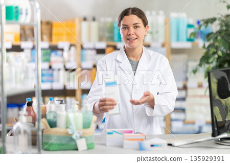 Skilled young woman pharmacist standing at prescription counter and inviting clients at pharmacy shop Skilled young woman pharmacist standing at prescription counter and inviting clients at pharmacy shop 135929391