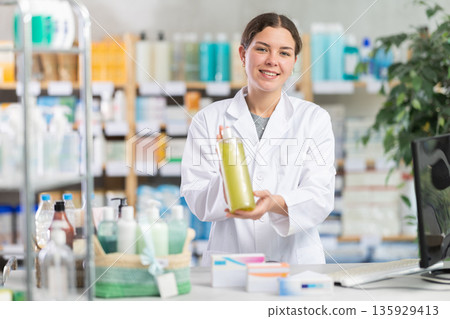 Skilled happy young woman pharmacist standing at prescription counter and offering shampoo or shower gel to clients at pharmacy Skilled happy young woman pharmacist standing at prescription counter and offering shampoo or shower gel to clients at pharmacy 135929413