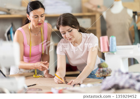 Manager of sewing workshop teaches a young girl seamstress to draw using patterns 135929452
