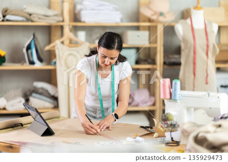Woman tailor draws dress pattern of product on paper, tablet on table. 135929473