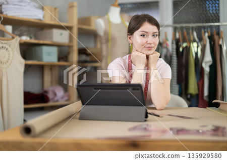 Seamstress girl draws outline of future dress on an electronic tablet in sewing workshop Seamstress girl draws outline of future dress on an electronic tablet in sewing workshop 135929580