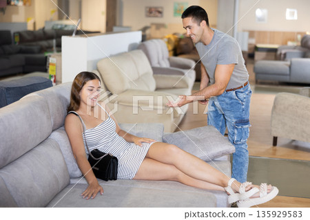Couple chooses furniture in store.Wife is sitting on sofa,husband is standing next to her Couple chooses furniture in store.Wife is sitting on sofa,husband is standing next to her 135929583