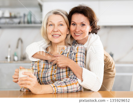 Old woman sitting at the kitchen table and middle-aged woman embracing her Old woman sitting at the kitchen table and middle-aged woman embracing her 135929652