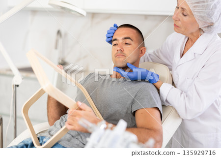 Portrait of man sitting and looking at mirror during appointment with doctor at cosmetology clinic 135929716