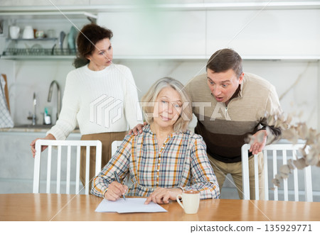 Old woman compiling heritage document while middle-aged man and woman competing in the kitchen Old woman compiling heritage document while middle-aged man and woman competing in the kitchen 135929771