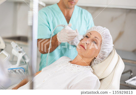 Cosmetologist performing anti-aging face hardware therapy procedure for aged female client in clinic of aesthetic medicine Cosmetologist performing anti-aging face hardware therapy procedure for aged female client in clinic of aesthetic medicine 135929813