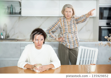 Upset middle-aged woman sitting at the kitchen table with her back to old woman quarreling to her Upset middle-aged woman sitting at the kitchen table with her back to old woman quarreling to her 135929852