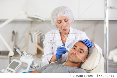 Female cosmetologist performing anti-aging face phototherapy procedure for male client in clinic of aesthetic clinic Female cosmetologist performing anti-aging face phototherapy procedure for male client in clinic of aesthetic clinic 135929855