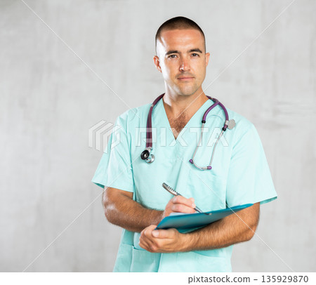 Portrait of male doctor in medical gown with stethoscope making notes in patient medical history Portrait of male doctor in medical gown with stethoscope making notes in patient medical history 135929870