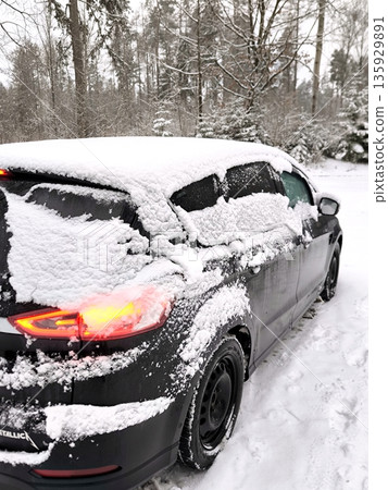 A black modern car covered in snow with its taillights on a winter road 135929891