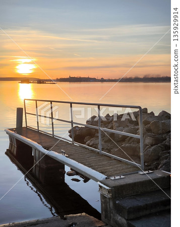 Wooden pier with railings on a rocky shore at sunset. Wooden pier with railings on a rocky shore at sunset. 135929914