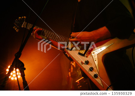 Close-up of a guitarists hands on a flying V electric guitar with microphone stand nearby, lit by warm orange stage lights and haze. Close-up of a guitarists hands on a flying V electric guitar with microphone stand nearby, lit by warm orange stage lights and haze. 135930329