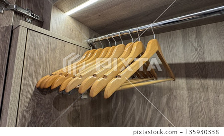Wooden clothes hangers hanging on a metal rod inside a wooden wardrobe, with an LED light strip above. 135930338