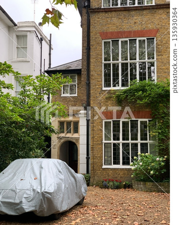 London brick townhouse facade with large white windows, ivy, arched entry, gravel yard and a car covered with a grey tarp. London brick townhouse facade with large white windows, ivy, arched entry, gravel yard and a car covered with a grey tarp. 135930364