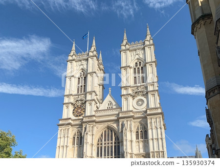 London view of Westminster Abbey west towers with clock, Gothic stone facade and a flagpole against a clear blue sky with clouds. 135930374