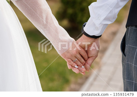 Close-up of a bride and groom holding hands during wedding ceremony, white lace sleeves and formal attire, soft natural lighting, romantic outdoor setting Close-up of a bride and groom holding hands during wedding ceremony, white lace sleeves and formal attire, soft natural lighting, romantic outdoor setting 135931479