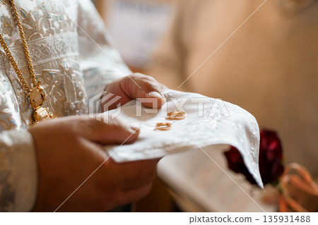 Wedding rings held in white cloth by priest during ceremony, gold bands on ornate vestments with red rose backdrop 135931488
