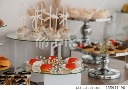Elegant dessert display with white chocolate-covered treats, red macarons, and baby's breath flowers on glass tiered stands Elegant dessert display with white chocolate-covered treats, red macarons, and baby's breath flowers on glass tiered stands 135931498