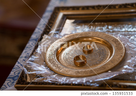 Golden Wedding Rings Resting on Ornate Religious Plate in Church Setting with Rich Textures and Warm Lighting 135931533