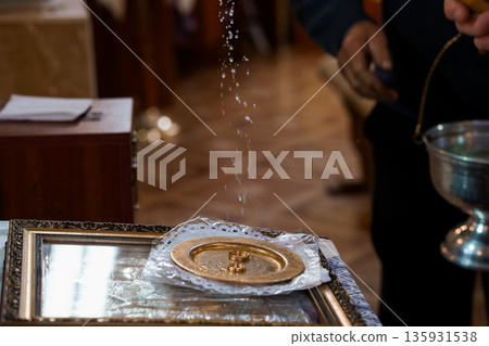 Priest Blessing Wedding Rings with Holy Water During Ceremony in Church Interior with Golden Plate and Ornate Frame 135931538
