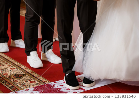 Elegant wedding ceremony feet close-up with bride in white gown and groom in black suit on red carpet with traditional embroidered runner 135931540