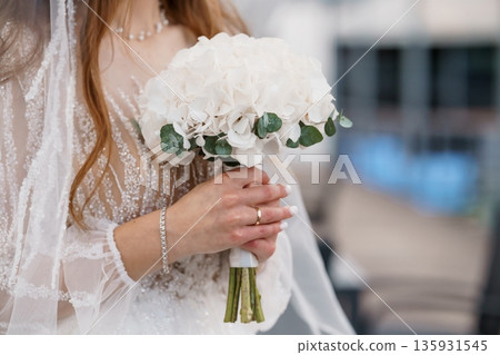 Elegant bride holding white hydrangea bouquet with green eucalyptus, lace wedding dress, pearl necklace, and veil in soft natural light Elegant bride holding white hydrangea bouquet with green eucalyptus, lace wedding dress, pearl necklace, and veil in soft natural light 135931545