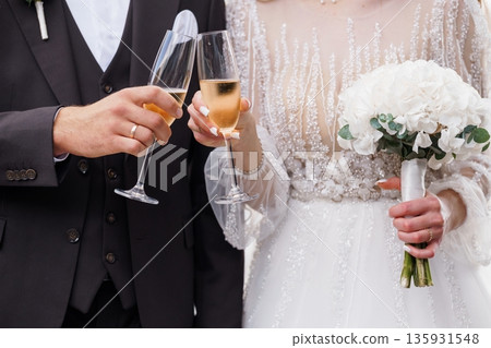 Bride and groom clinking champagne glasses in elegant wedding attire with white floral bouquet and sparkling dress details 135931548