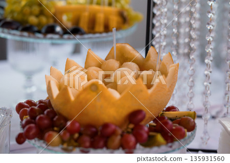 Elegant Fruit Display with Melon Carving and Grapes on Glass Stand for Party Catering and Event Decorations Elegant Fruit Display with Melon Carving and Grapes on Glass Stand for Party Catering and Event Decorations 135931560