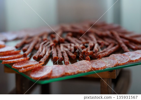 A Close-Up View of Sliced Salami and Stick-Style Salami on a Glass Tray with Warm Lighting and Rustic Background A Close-Up View of Sliced Salami and Stick-Style Salami on a Glass Tray with Warm Lighting and Rustic Background 135931567