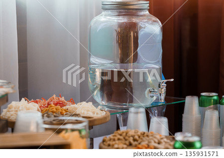 Large Glass Water Dispenser with Metal Faucet on Buffet Table Surrounded by Snacks and Drinks 135931571