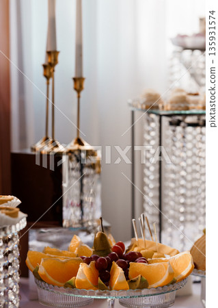 Elegant Fruit Platter Arrangement with Citrus Slices and Grapes on Glass Stand Amid Crystal Decor and Candlelight Ambiance 135931574