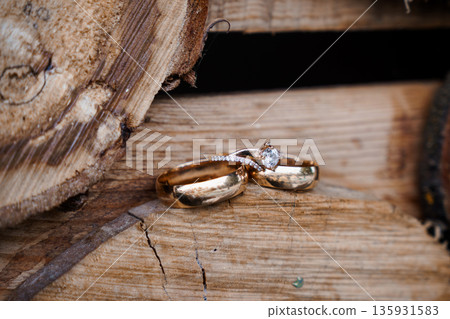 Golden wedding rings with diamond engagement ring resting on rustic wooden log surface, natural texture and warm tones 135931583